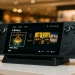Steam Deck OLED displayed on a retail demo stand in a well-lit department store, screen showing game titles like Elden Ring and Cyberpunk 2077