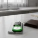 A small Apple AirTag tracker in a green circular holder attached to a car remote sitting on an iPhone and wallet on a marble counter.