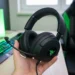 Hand holding a black Razer Kraken gaming headset with extended microphone and glowing RGB earcups against a bright gaming desk setup with green LED lighting.