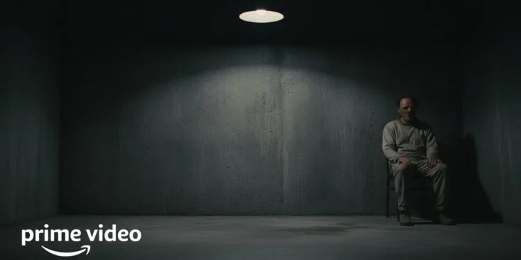 Figure in a restraint mask sits in a dark clinical cell under a spotlight.