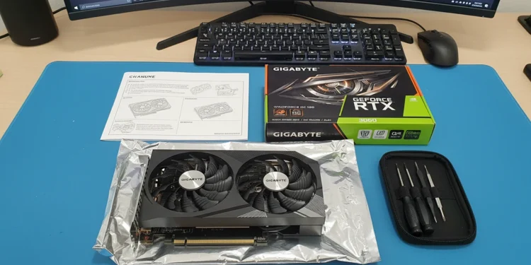 A Gigabyte GeForce RTX 3060 graphics card unboxed on a blue desk mat with its retail box, manual, and a small precision screwdriver kit.