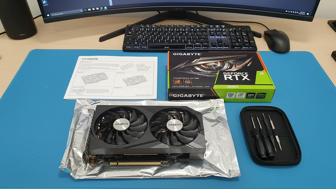 A Gigabyte GeForce RTX 3060 graphics card unboxed on a blue desk mat with its retail box, manual, and a small precision screwdriver kit.