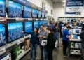 Shoppers comparing prices on new TVs and laptops in a crowded electronics store, illustrating tech trends to watch as gadget prices rise