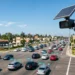 Flock license plate reader camera with solar panel mounted on a pole above a busy intersection, tracking passing vehicles